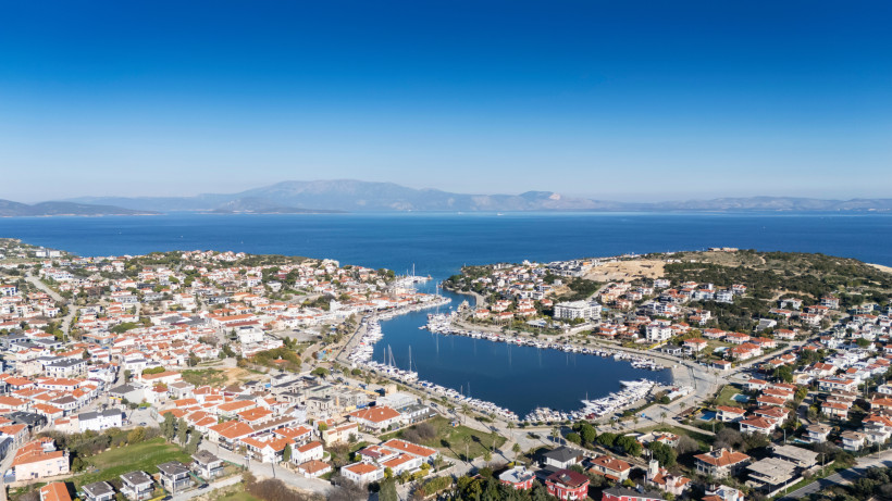 Panoramablick auf das Küstenstädtchen Çeşme an der türkischen Ägäis mit rotdachigen Häusern, einem Yachthafen in der Mitte und dem tiefblauen Meer im Hintergrund. Am Horizont sind sanfte Hügel und griechische Inseln zu sehen.