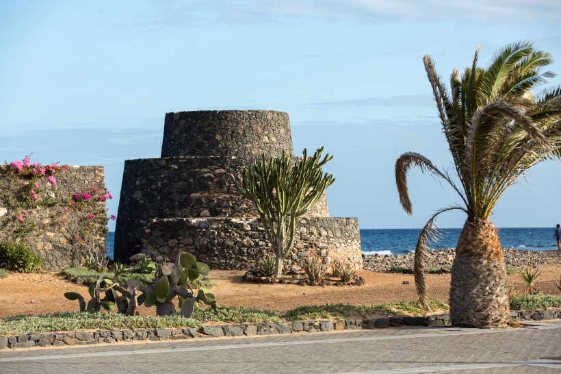 Castillo de San Buenaventura aus dunklem Naturstein an der Küste, mit Palmen und Sukkulenten im Vordergrund