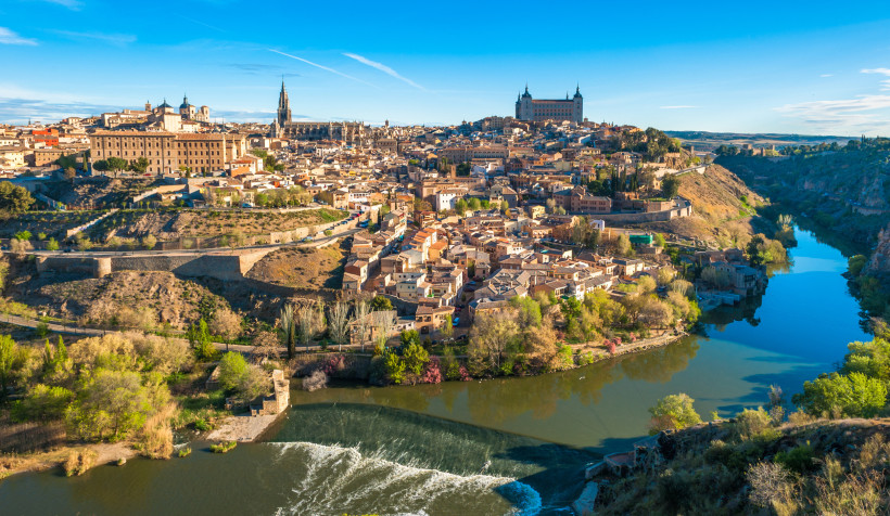 Panoramablick auf die Altstadt von Toledo, Spanien, umgeben vom Fluss Tajo. Gut sichtbar sind die Kathedrale von Toledo und der Alcázar. Die Stadt liegt malerisch auf einem Hügel, umgeben von grüner Landschaft.