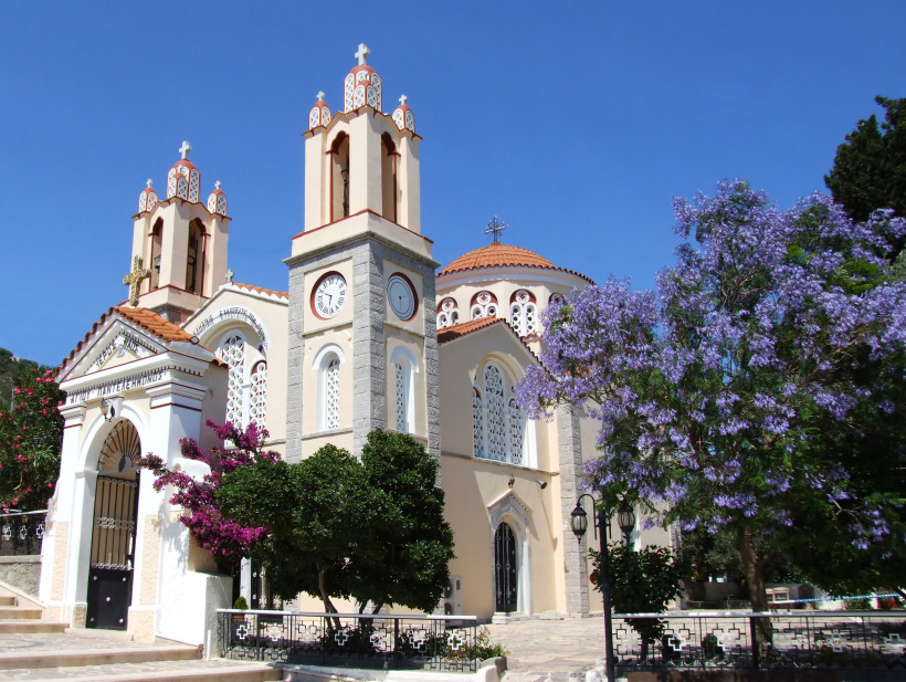 Griechisch-orthodoxe Kirche mit zwei Glockentürmen und einer Kuppel, verziert mit weißen Ornamenten und Kreuzen. Vor dem Gebäude stehen blühende Bougainvillea und ein Baum mit violetten Blüten. Die Fassade ist hell gestrichen, das Dach mit roten Ziegeln g