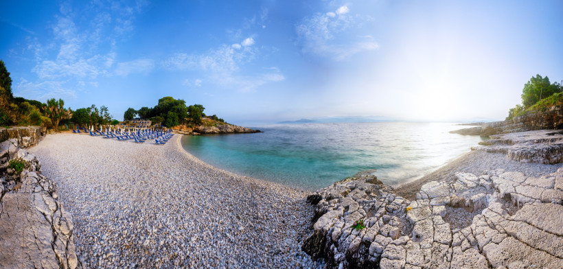 Korfu: Panoramablick auf den Kanoni Beach Panoramablick auf den Kiesstrand Kanoni auf Korfu mit Liegestühlen, türkisfarbenem Meer und Felsenküste