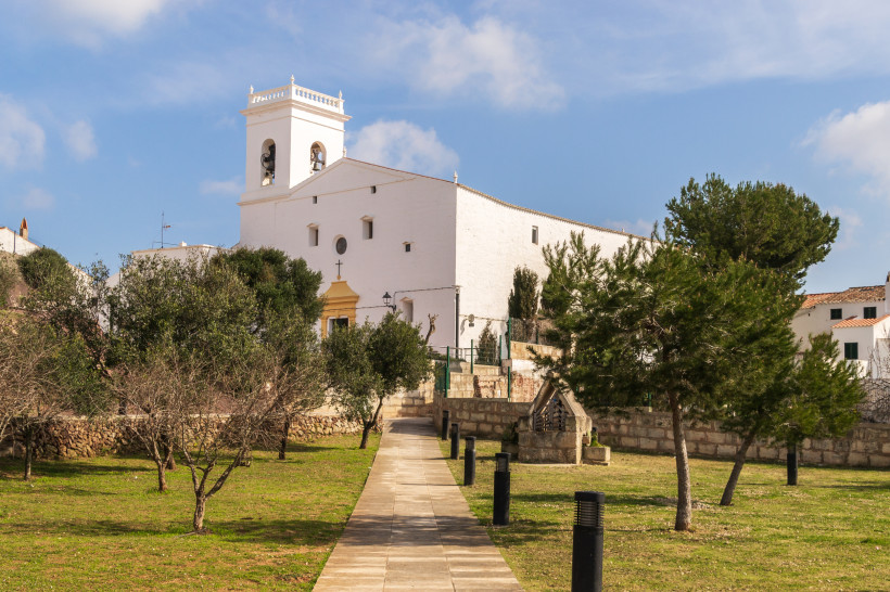 Menorca - Es Mercadal Weiß getünchte Kirche mit Glockenturm in Es Mercadal, Menorca. Ein gepflasterter Weg führt durch eine kleine Grünanlage mit Bäumen zur Kirche.