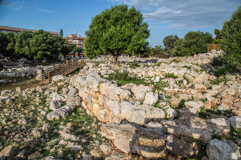 Steinruinen und niedrige Mauern der talaiotischen Siedlung von S’Illot mit Bäumen und Holzstegen
