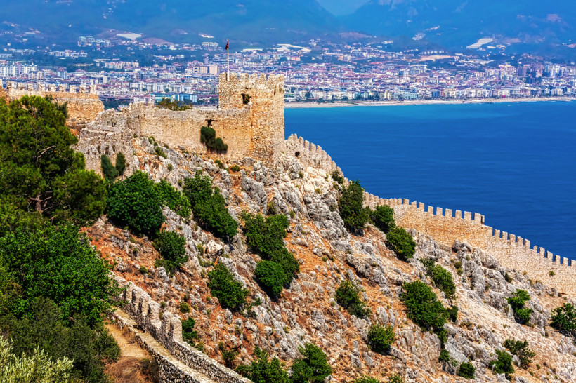 Detailansicht der alten Festungsmauern von Alanya auf einem felsigen Hügel mit üppigem Grün, im Hintergrund die Küstenstadt und das blaue Meer