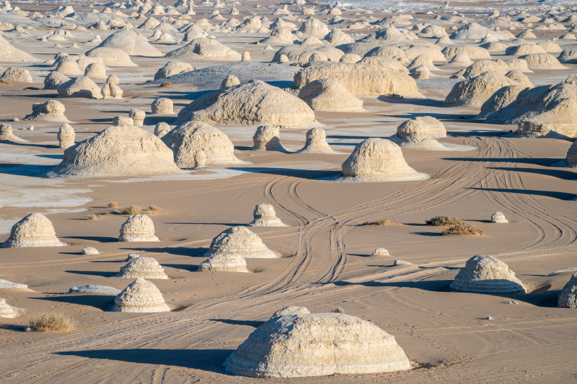 Das Bild zeigt eine außergewöhnliche Landschaft im White Desert Nationalpark in Ägypten. Die sandige Ebene ist von zahlreichen hellen Kalksteinformationen übersät, die wie riesige Pilze, Hügel oder Kuppeln geformt sind. Der Untergrund wechselt zwischen we
