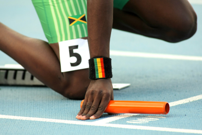 Jamaika Ein jamaikanischer Leichtathlet in Startposition für ein Staffelrennen auf einer blauen Laufbahn. Der Sportler trägt grüne Laufkleidung mit der Flagge Jamaikas und die Startnummer 5. Am Handgelenk trägt er ein schwarzes Schweißband mit den Farben Rot, Gel