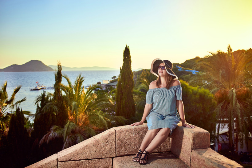 Ferienzene an der türkischen Ägäis mit Frau auf einer Aussichtsterrasse, Blick auf Meer, Palmen, Küste und mediterrane Landschaft