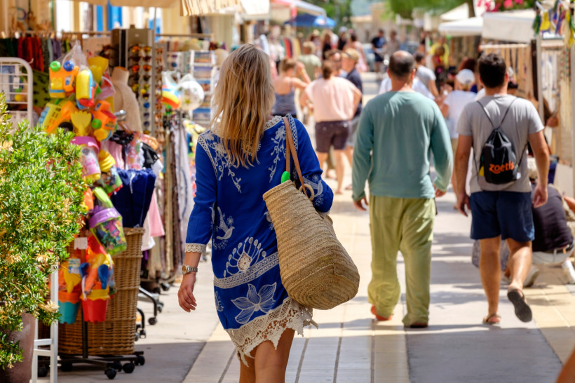 Formentera Touristin mit blauem Kleid und Strohtasche geht über einen belebten Markt. Um sie herum sind viele Menschen und bunte Stände mit Kleidung, Spielzeug und Souvenirs.