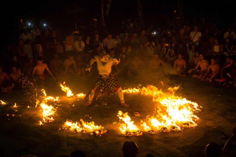 Bali Ein traditioneller balinesischer Tänzer mit weißer Körperbemalung, Maske und kariertem Sarong führt mitten in einem brennenden Kreis einen dramatischen Feuertanz auf. Rundherum sitzen Zuschauer im Dunkeln, viele mit gezückten Handys. Das Bild zeigt eine S