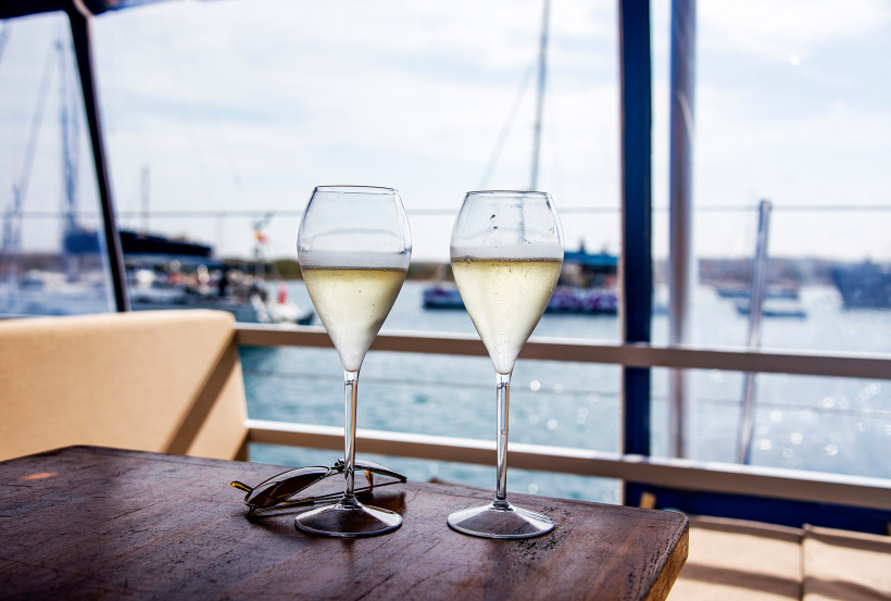 Zwei gefüllte Weingläser mit gekühltem Weißwein stehen auf einem Holztisch in einem Hafenrestaurant. Im Hintergrund verschwimmen Segelboote und Wasser im Sonnenlicht. Eine Sonnenbrille liegt neben den Gläsern auf dem Tisch.
