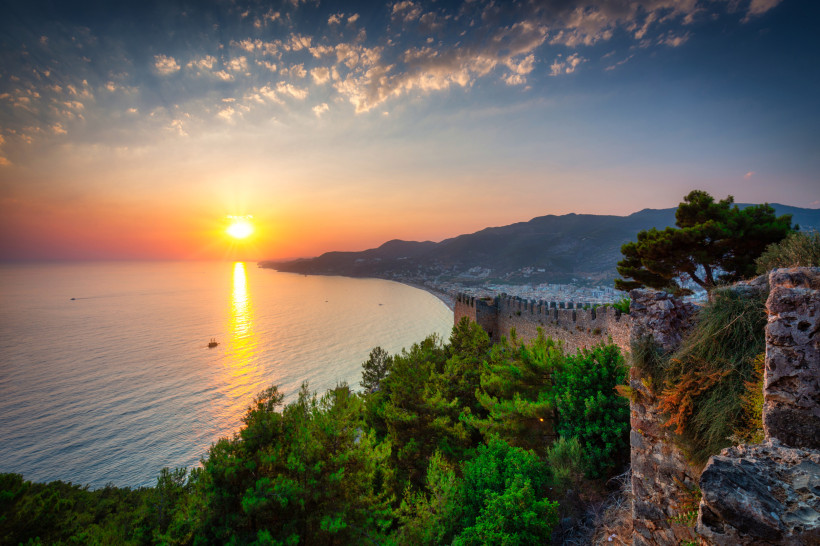 Das Bild zeigt die steinernen Mauern der historischen Festung von Alanya im Vordergrund. Im Hintergrund geht die Sonne über dem ruhigen Mittelmeer unter, während die Umgebung von dichtem Wald und Hügeln umgeben ist.