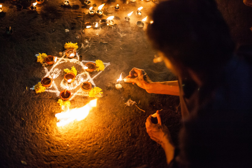 tief spirituelle Szene während eines hinduistischen Festes, vermutlich Diwali, dem Lichterfest. Eine Person ist dabei, eine kleine Öllampe (Diya) anzuzünden, umgeben von vielen weiteren brennenden Lichtern. Der Boden ist kunstvoll mit weißen Mustern aus R