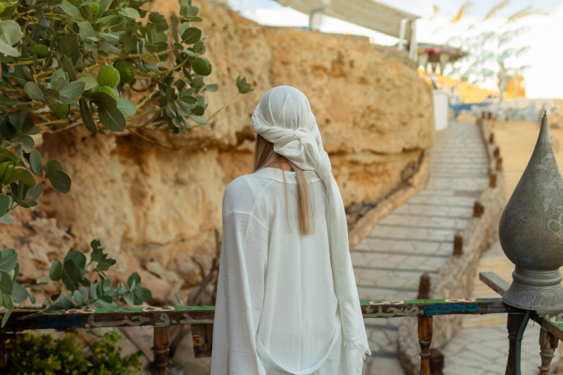 Ägypten - Sharm el Sheikh Eine Frau mit langen blonden Haaren steht mit dem Rücken zur Kamera und trägt ein weißes, locker fallendes Gewand und ein Tuch über dem Kopf. Sie blickt auf einen felsigen Weg mit Treppen, der sich entlang einer Steinwand durch eine Wüstenlandschaft schlä