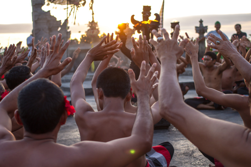 Bali Mehrere balinesische Männer sitzen im Halbkreis und strecken synchron die Arme in die Höhe – Teil einer traditionellen Kecak-Aufführung im Freien. Die Tänzer tragen rot-schwarz-karierte Sarongs und rote Blumen hinter den Ohren. Im Hintergrund versinkt die