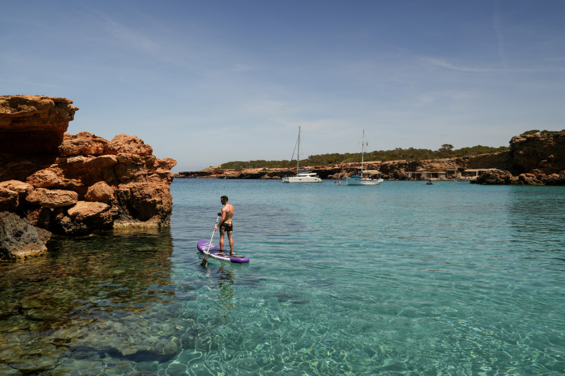 Ibiza Ein Mann steht auf einem lilafarbenen Stand-Up-Paddleboard auf glasklarem, türkisblauem Wasser. Im Hintergrund liegen Segelboote in einer ruhigen Bucht, umgeben von Felsen und niedriger Vegetation. Der Himmel ist wolkenlos, eine Flugzeugspur zieht sich üb