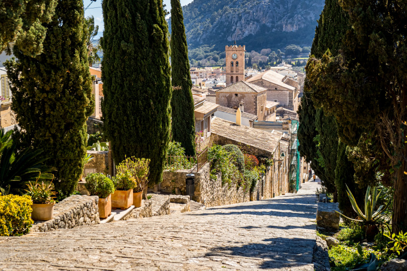 eine Steinstraße unten in der Stadt Pollensa, umgeben von Bäumen und mit Blick auf die Berge