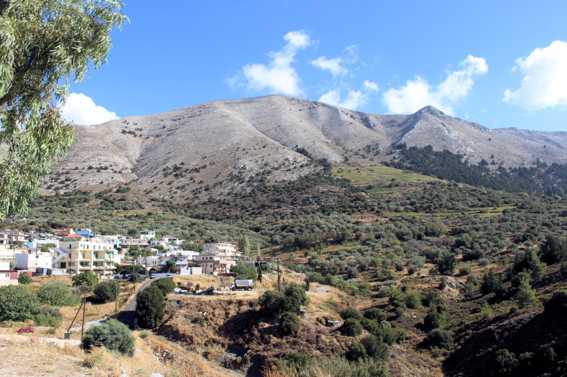 Das Bild zeigt ein ländliches Dorf ( Embona ) am Fuße eines felsigen Gebirges auf einer griechischen Insel, vermutlich Kreta.   Die Häuser sind meist weiß oder hellbeige mit Flachdächern und liegen eingebettet zwischen sanften Hügeln und Olivenhainen.   I