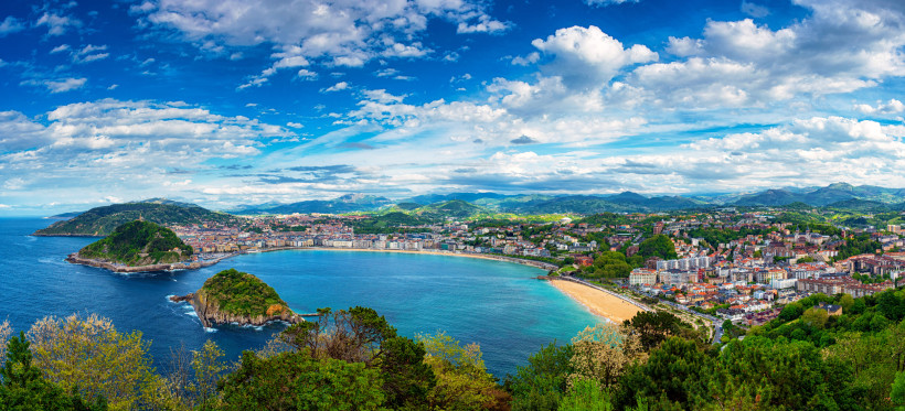 Panoramablick auf die Bucht La Concha in San Sebastián mit Strand, Stadt und grünen Hügeln unter wolkigem Himmel