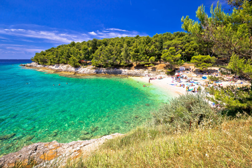 Malerische Badebucht bei Pula in Kroatien mit türkisfarbenem Wasser, hellem Kiesstrand und vielen Badegästen. Im Hintergrund dichter Pinienwald, der bis an das Ufer reicht. Der Himmel ist strahlend blau, einige Menschen schwimmen im Meer oder entspannen a