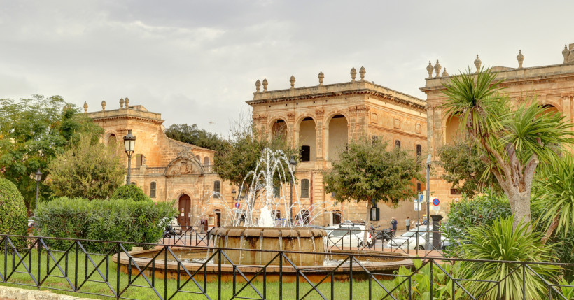 Menorca - Ciutadella de Menorca Historische Gebäude aus Sandstein an der Plaça des Born in Ciutadella de Menorca mit Springbrunnen im Vordergrund und Palmen im Park.