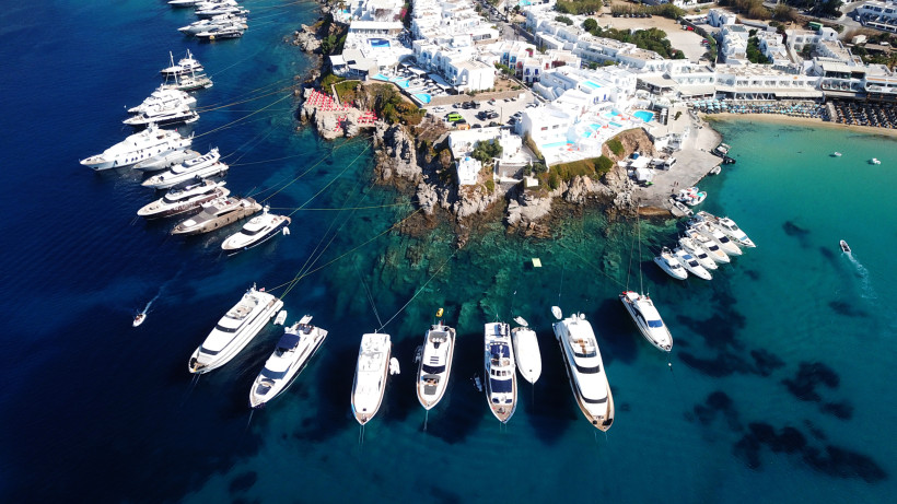 Luftaufnahme einer kleinen Halbinsel mit weißen Häusern und Pools, umgeben von türkisfarbenem Wasser. Zahlreiche große Yachten und Boote sind entlang der Küste vertäut. Die Szene zeigt klares Wasser und felsige Küstenlinien