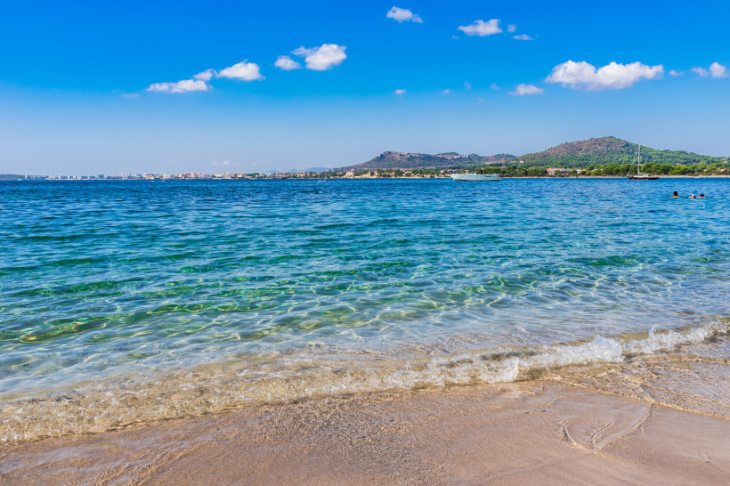 Flacher Sandstrand mit klarem, türkisfarbenem Wasser bei Cala Bona auf Mallorca, kleine Boote vor der Küste und Blick entlang der Bucht Richtung Costa dels Pins.