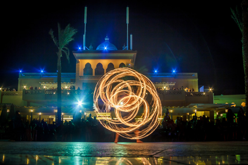 Ägypten Feuershow bei Nacht: Ein Künstler wirbelt brennende Fackeln, sodass kreisförmige Lichtspuren entstehen. Im Hintergrund ein orientalisches Hotel mit beleuchteten Balkonen und Publikum.