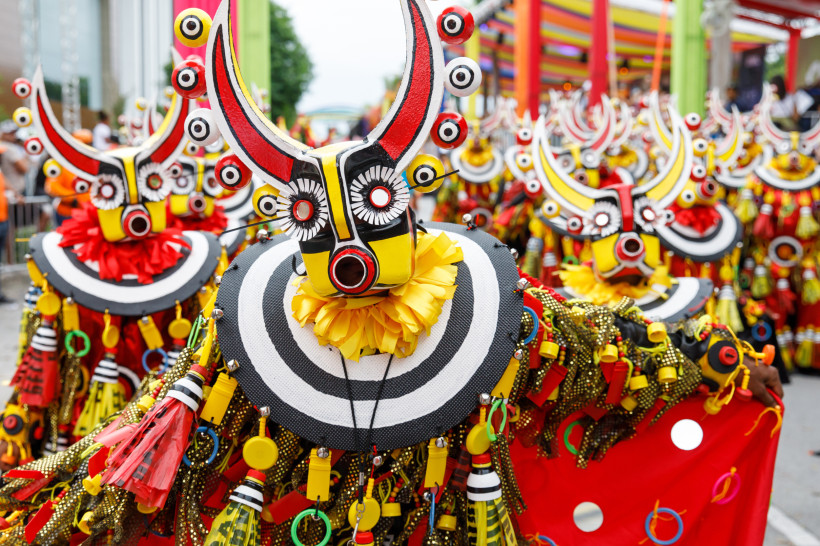 Teilnehmer eines Karnevalsumzugs mit auffälligen Masken in Rot, Gelb und Schwarz, verziert mit Hörnern, Kreisen und Fransen, in einer bunten Straßenparade