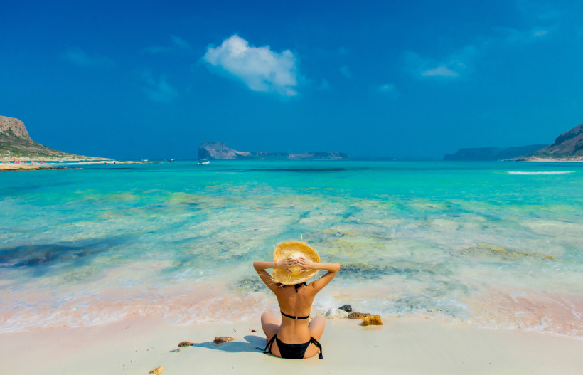 Balos-Lagune mit türkisfarbenem, flachem Wasser und Sandstrand; Person mit Strohhut sitzt am Ufer und blickt aufs Meer