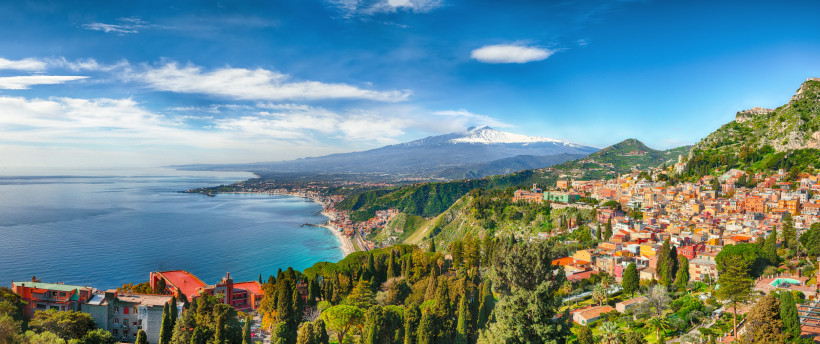 Panoramablick auf die sizilianische Küstenstadt Taormina, die sich terrassenförmig an einen grünen Berghang schmiegt. Rechts im Bild die farbenfrohen Gebäude der Altstadt, mittig der grüne Bergrücken, im Hintergrund der schneebedeckte Vulkan Ätna. Links d