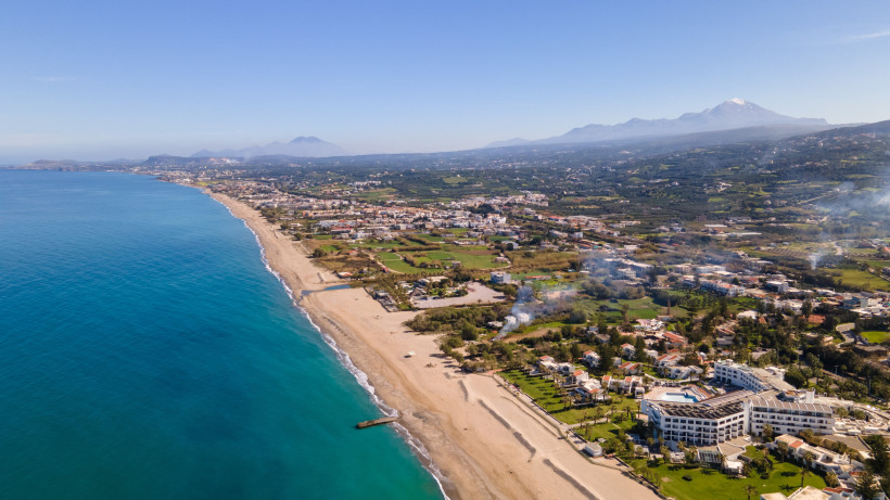 Luftaufnahme des Strandes von Rethymno mit Küstenlinie, Bebauung und Bergen im Hintergrund