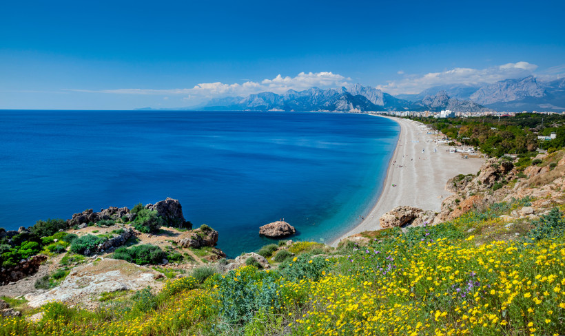 Konyaalti Beach Panorama – Türkisblaues Meer und Taurusgebirge in Antalya Panoramablick auf den Konyaalti Beach in Antalya mit tiefblauem Meer, Kiesstrand, blühender Küstenlandschaft und beeindruckendem Taurusgebirge im Hintergrund.