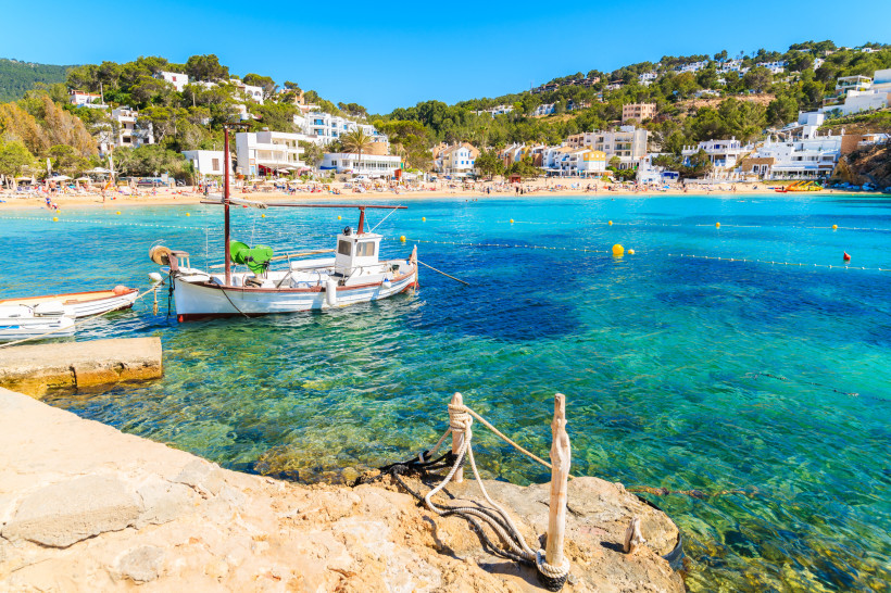 Ibiza Ein traditionelles weißes Fischerboot mit rotem Mast liegt in einer türkisblauen Bucht auf Ibiza vor Anker. Im Hintergrund erstreckt sich ein belebter Sandstrand mit vielen Menschen, dahinter liegen weiße und pastellfarbene Häuser, eingebettet in grüne Hü