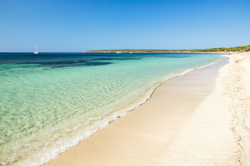 Weitläufiger Sandstrand der Playa de Migjorn auf Formentera mit flach abfallendem, glasklarem Wasser. Im Hintergrund segeln Boote auf dem tiefblauen Meer.