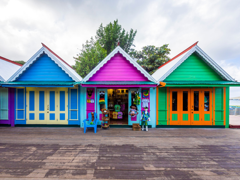 Jamaika Drei kleine, bunte Holzhäuser im karibischen Stil nebeneinander. Das linke Haus ist blau mit gelben Türen, das mittlere pink-lila mit einem geöffneten Souvenirshop und das rechte grün mit orangefarbenen Türen. Vor dem mittleren Laden stehen bunte Figuren,