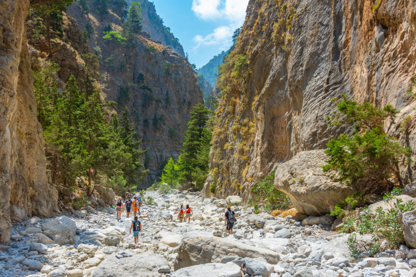 Kreta Eine Gruppe von Wanderern durchquert eine beeindruckende Schlucht mit hohen, steilen Felswänden und grünen Kiefern. Die Landschaft ist felsig, und der Weg führt über große Steine. Der Himmel ist blau mit einigen Wolken, die Sonne scheint hell.