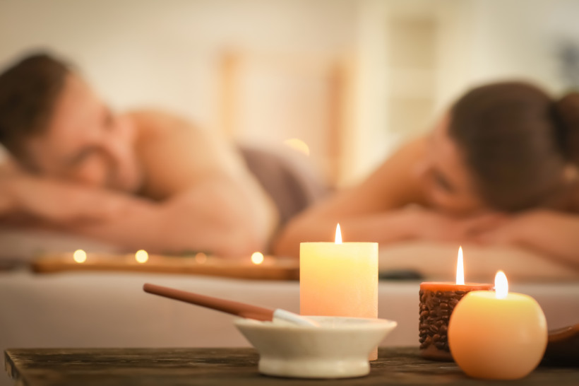 Spa-Komposition mit Kerzen und einem entspannten Paar im Hintergrund Paar liegt entspannt auf Massageliegen in einem Spa, mit brennenden Kerzen und Räucherstäbchen im Vordergrund, in ruhiger und romantischer Atmosphäre.