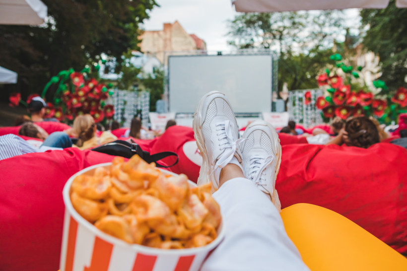 Blick aus der Perspektive einer liegenden Person in einem Open-Air-Kino. Im Vordergrund weiße Sneakers, daneben ein großer Eimer mit Chips. Viele Zuschauer liegen entspannt auf roten Sitzsäcken vor einer großen Leinwand. Die Kulisse ist von Bäumen und dek