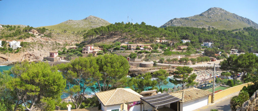 Panorama der Cala Molins in Cala San Vicente mit türkisfarbener Bucht, Hanghäusern und Bergen der Serra de Tramuntana im Hintergrund.