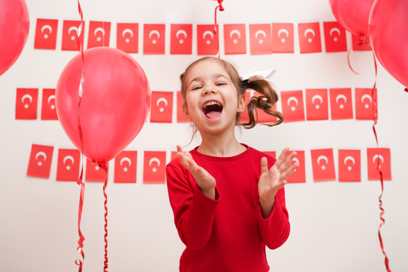 Ein lachendes Mädchen in rotem Pullover steht vor einer Wand mit türkischen Flaggen und roten Luftballons. Sie klatscht in die Hände und wirkt fröhlich und ausgelassen.