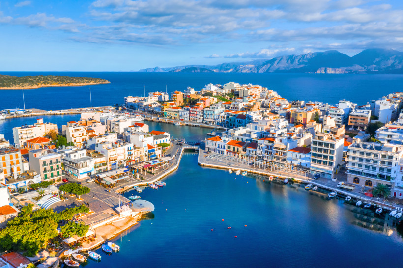 Blick auf den Hafen von Agios Nikolaos mit Booten, Promenade und dicht bebauter Küstenstadt vor blauem Meer und Bergen