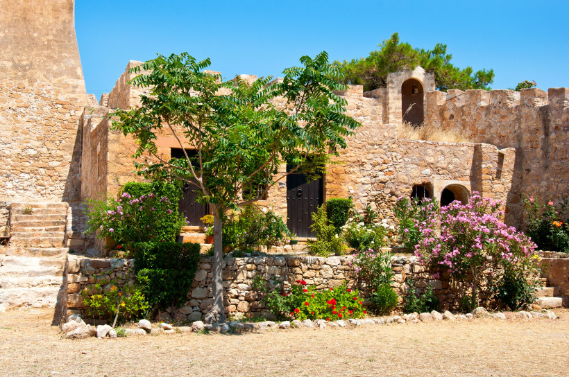 Historisches mediterranes Steingebäude mit Rundbögen, schwarzen Holztüren und blühenden Pflanzen wie Oleander und Rosen. Ein kleiner Baum und gepflegte Büsche schmücken den sonnigen Hof unter klarem, blauem Himmel