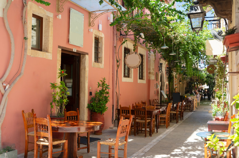 Altstadtgasse in Rethymno mit Straßencafé und Weinranken Gasse in der Altstadt von Rethymno mit Tischen und Stühlen vor einem rosafarbenen Gebäude unter Weinranken