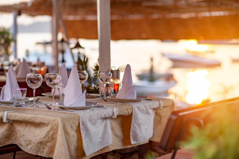 Gedeckter Tisch mit Weingläsern und gefalteten Servietten auf einer Terrasse am Wasser bei Sonnenuntergang