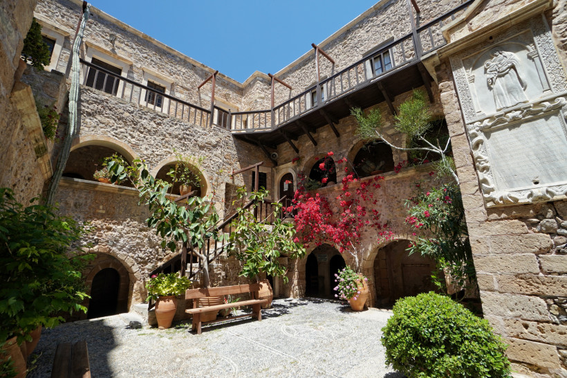 Traditioneller Kloster-Innenhof mit Steinbögen, Holzbalkonen und reich bepflanzten Blumentöpfen. Bougainvillea und Oleander ranken an den Mauern, eine Bank steht im Schatten