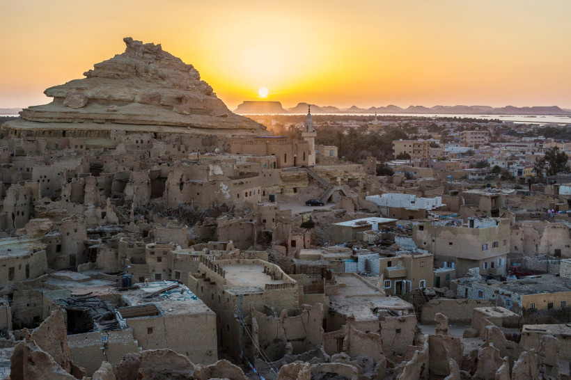 Das Bild zeigt die beeindruckende Lehmbau-Altstadt Schali in der Siwa-Oase, Westägypten, kurz vor Sonnenuntergang. Im Vordergrund erheben sich die verwitterten Überreste der traditionellen Häuser, die aus Salz, Lehm und Palmholz errichtet wurden. Der mark