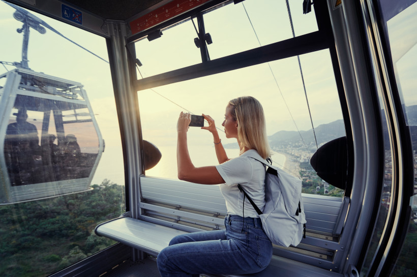 Eine junge Frau sitzt in einer Seilbahnkabine in Alanya und fotografiert den Ausblick auf das Meer und den Strand bei Sonnenuntergang.