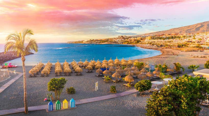 Blick über eine Strandbucht mit Strohschirmen, Promenade und Küstenstadt bei Sonnenuntergang Strand mit Reihen aus Strohschirmen und Liegen an einer Bucht bei Sonnenuntergang, im Hintergrund Küstenstadt und Berge