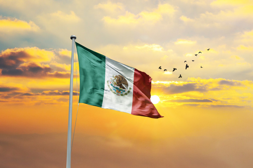 Unabhängigkeitstag Die mexikanische Flagge mit grün, weiß und rot weht im Wind vor einem goldenen Sonnenuntergang, während Vögel am Himmel fliegen