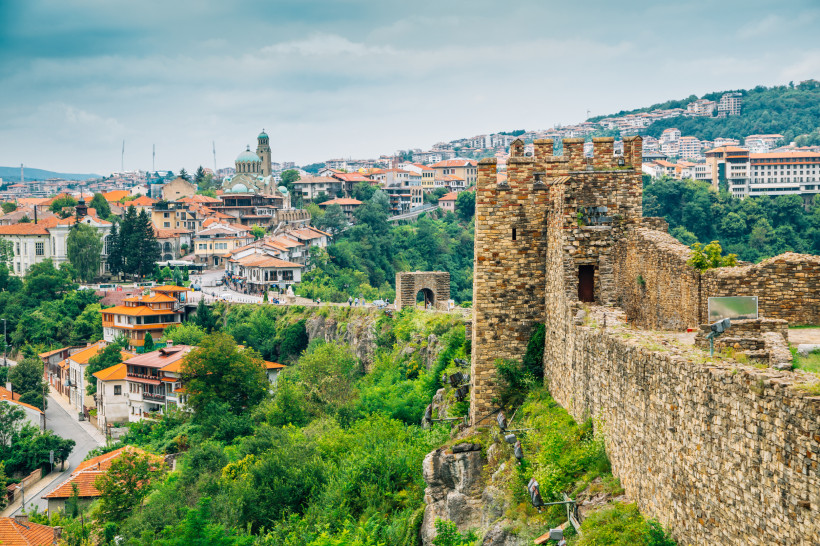 Blick auf die Altstadt von Veliko Tarnovo in Bulgarien. Rechts im Bild verläuft eine mittelalterliche Steinmauer mit einem Wachturm, links sind rote Ziegeldächer und verwinkelte Häuser auf dem grünen Hang verteilt. Im Hintergrund erhebt sich die markante 
